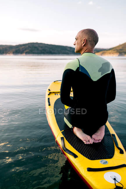 Вид сзади на взрослого мужчину в гидрокостюме, стоящего на коленях на гребной доске на спокойном озере — стоковое фото
