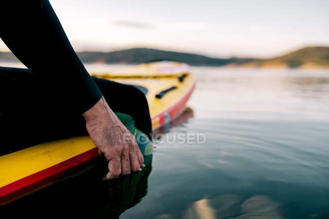 Crop unrecognizable male surfer sitting on SUP board and touching sea water at sunset — Stock Photo
