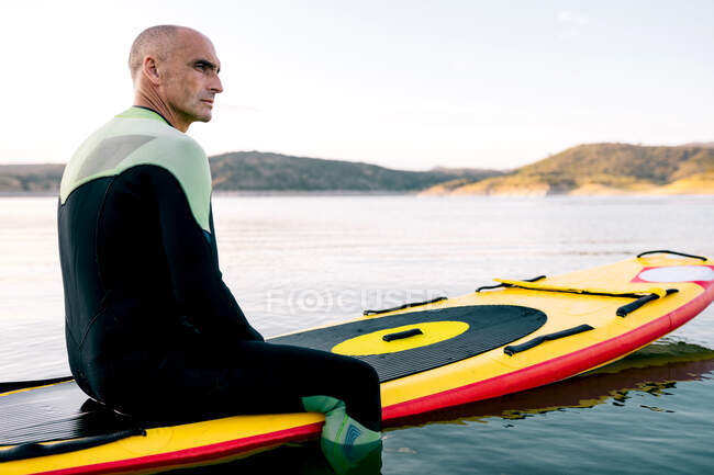 Side view of thoughtful adult male in wetsuit sitting on paddle board on calm lake water surface — Stock Photo