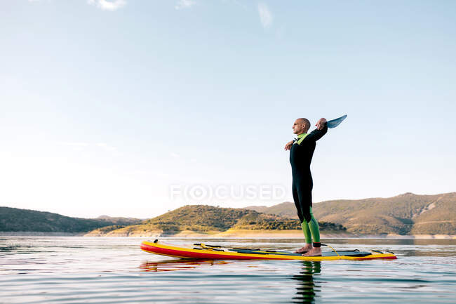 Вид сбоку серфингиста-мужчины с насадкой, стоящего на доске SUP в спокойном море и наслаждающегося закатом — стоковое фото