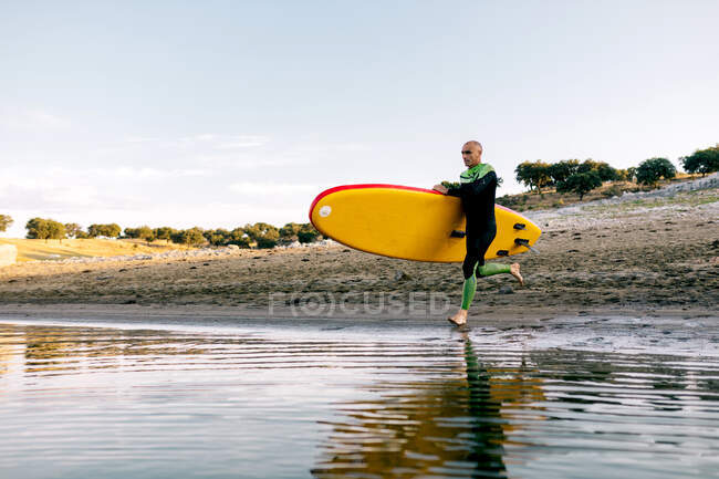 Full body of active sporty male in wetsuit carrying paddle board and entering river water — Stock Photo