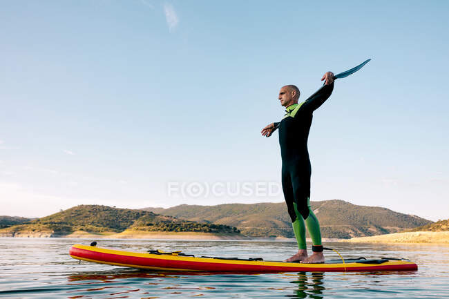 Vista lateral del sereno surfista masculino con la paleta de pie mirando hacia otro lado en la tabla SUP en el mar tranquilo y disfrutando de la puesta de sol - foto de stock