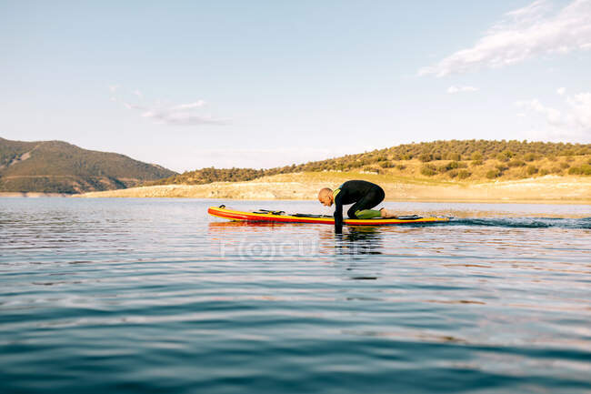 Ganzkörper-Seitenansicht eines erwachsenen Männchens im Neoprenanzug, das auf einem Paddelbrett kniet und mit den Armen auf einer ruhigen Wasseroberfläche paddelt — Stockfoto