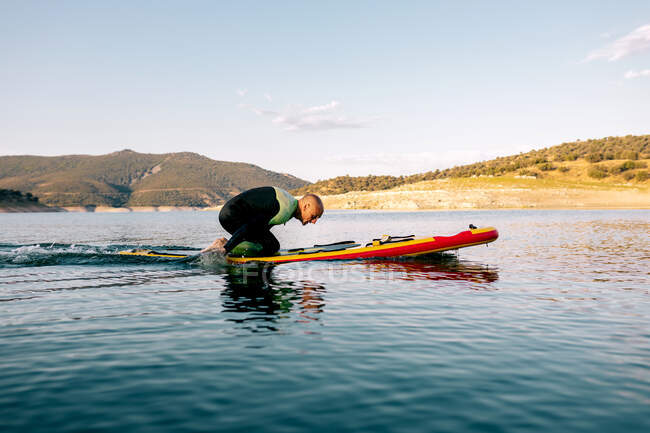 Vista lateral de cuerpo completo del macho adulto en traje de neopreno arrodillado sobre tabla de paddle y remando con los brazos en la tranquila superficie del agua del lago - foto de stock