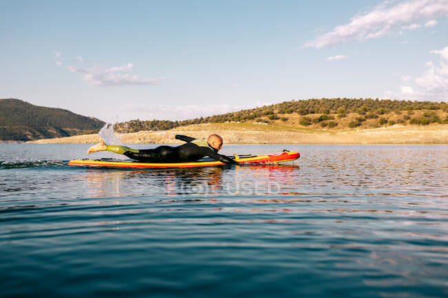 Vista laterale del maschio scalzo in muta sdraiato su pedana e nuoto sulla superficie del lago durante la pratica di sport acquatici durante la giornata estiva — Foto stock