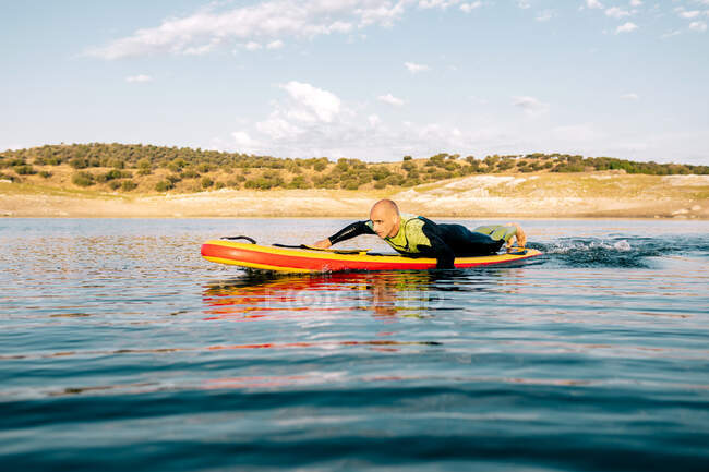 Seitenansicht eines barfüßigen Mannes im Neoprenanzug, der auf einem Paddelbrett liegt und auf der Oberfläche des Sees schwimmt, während er im Sommer Wassersport treibt — Stockfoto