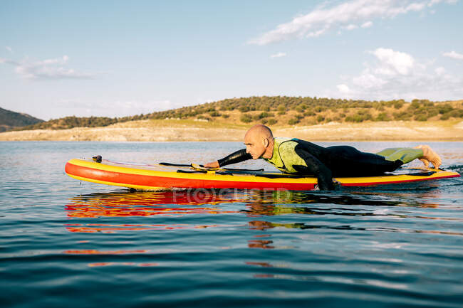 Vista laterale del maschio scalzo in muta sdraiato su pedana e nuoto sulla superficie del lago durante la pratica di sport acquatici durante la giornata estiva — Foto stock
