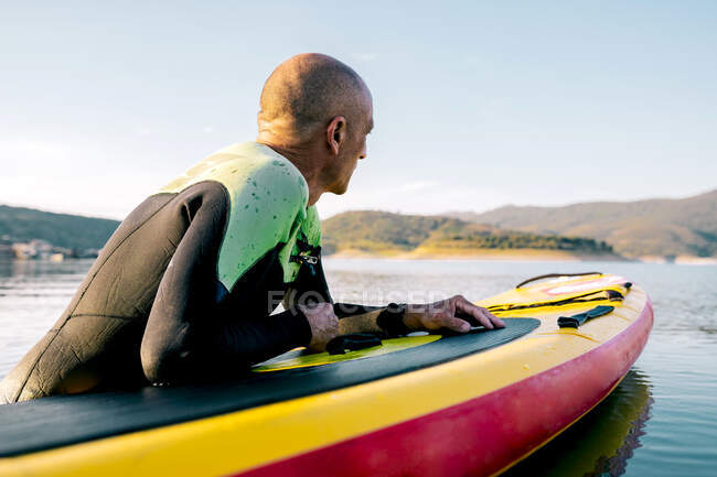 Вид збоку чоловіка в гідрокостюмі лежить на дошці весла і плаває на поверхні озера, займаючись водними видами спорту в літній день — стокове фото