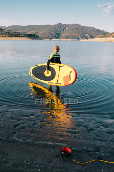 Vista lateral do surfista masculino em roupa de mergulho em pé com placa SUP amarela e remo na água do mar — Fotografia de Stock