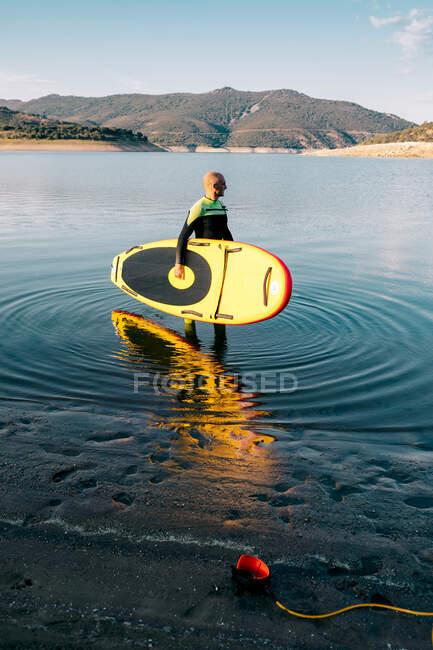 Vista lateral del surfista masculino en traje de neopreno de pie con tabla SUP amarilla y paleta en agua de mar - foto de stock