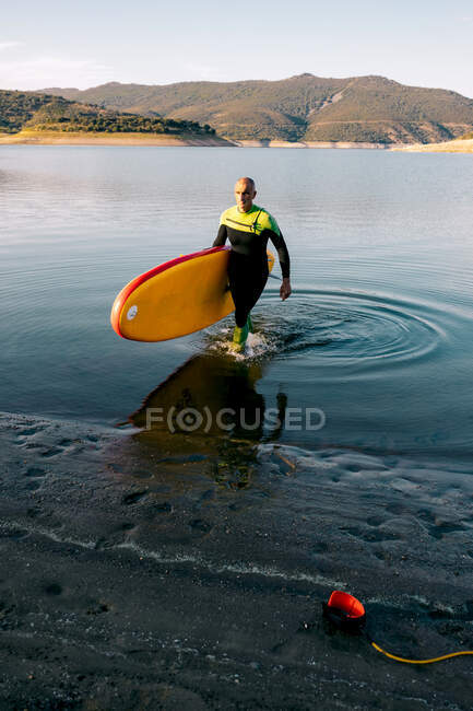 Surfeur masculin en combinaison de plongée marchant avec SUP board jaune et pagaie sur une plage de sable près de l'eau de mer — Photo de stock