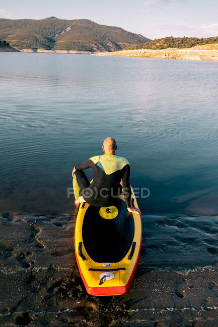 Back view of unrecognizable thoughtful male in wetsuit sitting on paddle board while preparing for rowing in lake water — Stock Photo