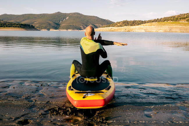 Вид ззаду на невпізнаваного чоловіка в гідрокостюмі, що сидить на дошці весла і розтягує руки і плечі, готуючись до веслування в озерній воді — стокове фото