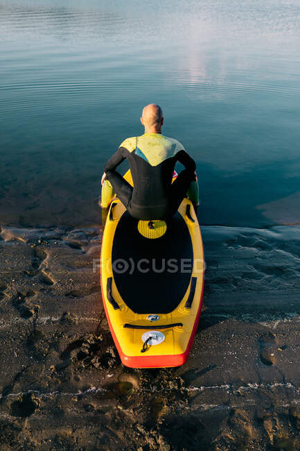 Задний вид неузнаваемого вдумчивого мужчины в гидрокостюме, сидящего на веслах во время подготовки к гребле в озере — стоковое фото
