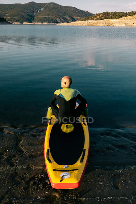 Rückenansicht eines unkenntlich gemachten Männchens im Neoprenanzug, das auf einem Paddelbrett sitzt, während es sich auf das Rudern im Seewasser vorbereitet — Stockfoto