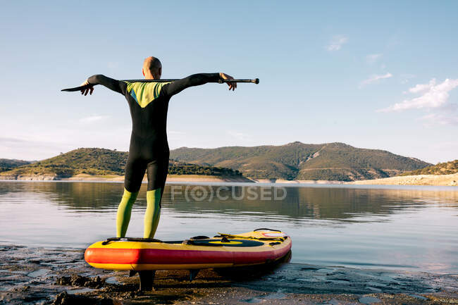Visão traseira do surfista masculino sereno com remo de pé olhando para longe na placa SUP na praia de areia perto do mar calmo e desfrutando de vistas — Fotografia de Stock