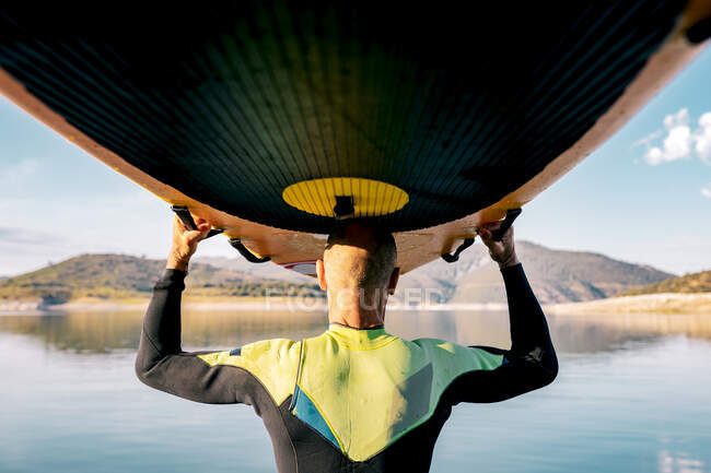 Vue arrière d'un surfeur masculin mature méconnaissable debout avec une planche à pagaie au-dessus de la tête près de la mer le soir et regardant ailleurs — Photo de stock