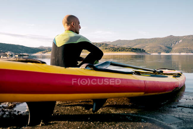 Back view of serene thoughtful male surfer with paddle sitting looking away on SUP board in sandy beach near calm sea and enjoying views — Stock Photo