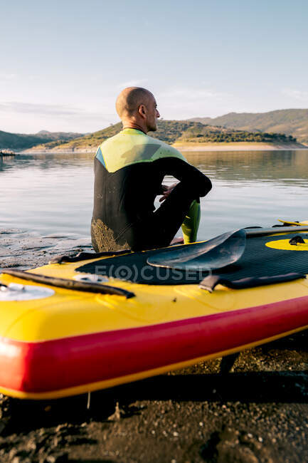 Back view of serene thoughtful male surfer with paddle sitting looking away on SUP board in sandy beach near calm sea and enjoying views — Stock Photo