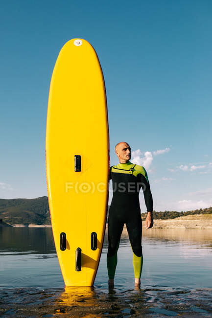 Чоловічий серфер у гідрокостюмі стоїть з жовтою дошкою СУП та веслою на піщаному пляжі біля морської води — стокове фото