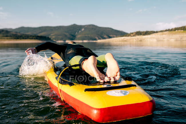 Back view of unrecognizable barefoot male in wetsuit lying on paddle board and swimming on lake surface while practicing water sport in summer day — Stock Photo