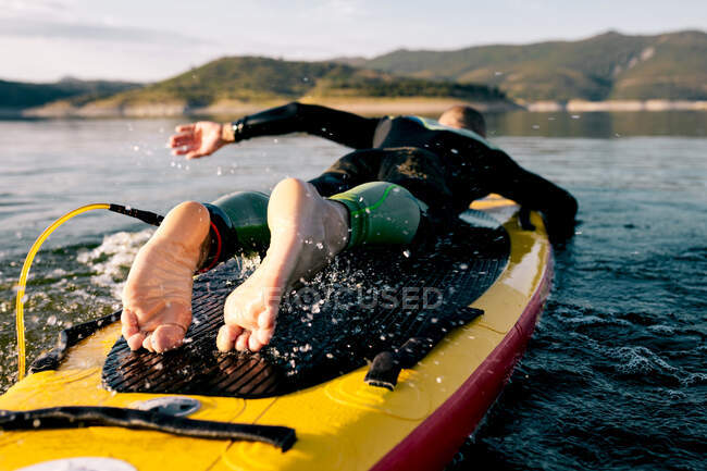 Vista posteriore del maschio scalzo irriconoscibile in muta sdraiato su pedana e che nuota sulla superficie del lago mentre pratica sport acquatici durante la giornata estiva — Foto stock