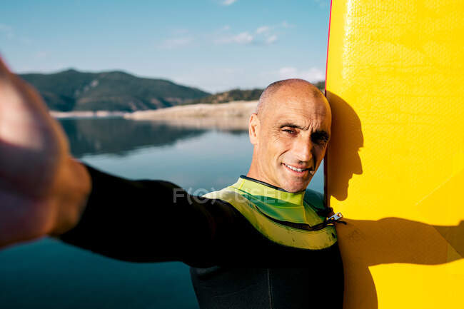 Hombre positivo de mediana edad activo en traje de neopreno de pie cerca de la tabla de paddle y mirando a la cámara mientras toma autorretrato contra el lago tranquilo en el día de verano - foto de stock