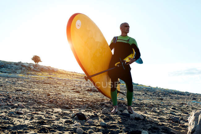 Niedrige Winkel Ganzkörper von selbstbewussten reifen Mann in Aktivkleidung tragen gelbes Paddelbrett und schauen weg, während sie am Sandstrand stehen — Stockfoto