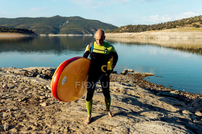 Male surfer in wetsuit walking with pump and SUP board on seashore and after surfing — Stock Photo