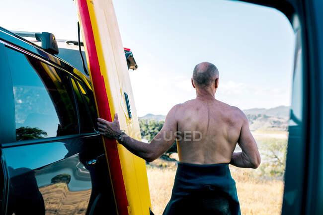 Back view of active middle aged shirtless man taking paddle board from automobile parked on lake shore in hilly nature while preparing for water sport practice in summer day — Stock Photo