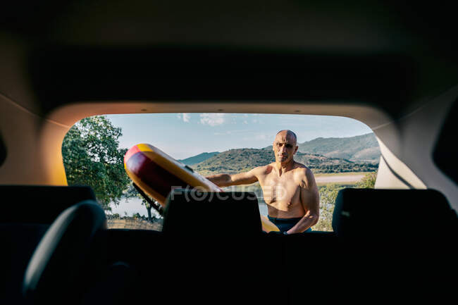 Aktiver Mann mittleren Alters ohne Hemd nimmt Paddelbrett aus Auto, das am Seeufer in hügeliger Natur geparkt ist, während er sich auf Wassersportübungen im Sommer vorbereitet — Stockfoto