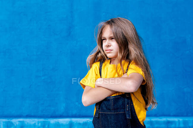 Adolescente segura de pie con los brazos cruzados sobre fondo azul y mirando hacia otro lado - foto de stock