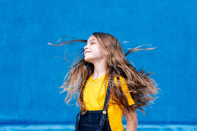 Entzückter verträumter Teenager mit langem fliegendem Haar, der mit geschlossenen Augen auf blauem Hintergrund steht — Stockfoto