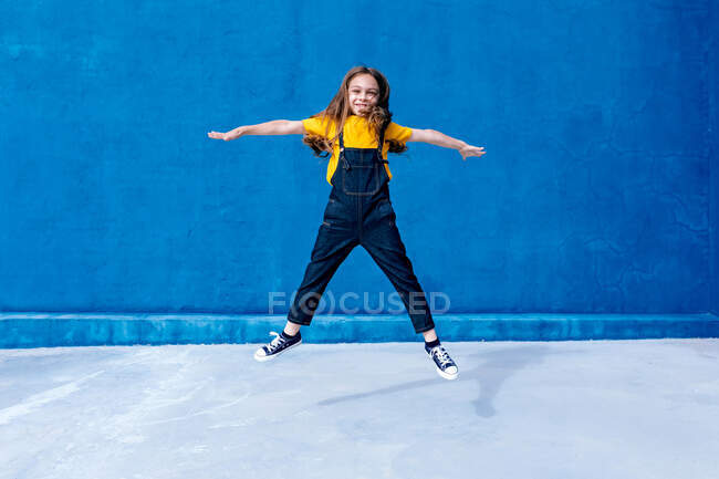 Carefree smiling teenager in moment of jumping above ground with outstretched arms on blue background — Stock Photo