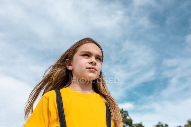 De baixo de adolescente sereno com cabelo longo e em roupa legal em pé no fundo do céu nublado azul e olhando para longe — Fotografia de Stock