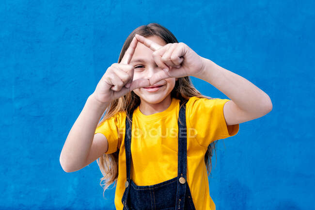 Adolescente alegre olhando através do buraco do sinal de mão triângulo no fundo azul no estúdio — Fotografia de Stock
