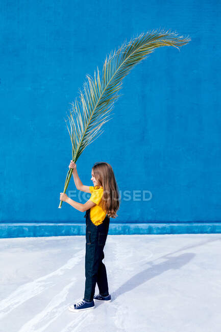 Vista lateral de adolescente de pie con enorme hoja de palmera en el fondo de la pared azul - foto de stock