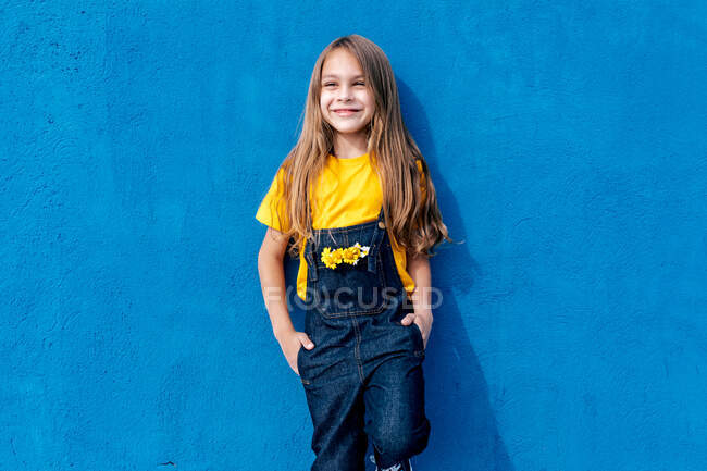 Cheerful teen hipster with bunch of yellow flowers in pocket of denim overall leaning on blue wall and looking away — Stock Photo