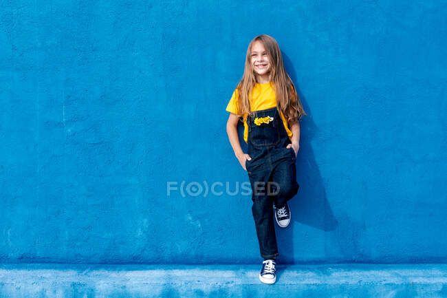 Cheerful teen hipster with bunch of yellow flowers in pocket of denim overall leaning on blue wall and looking away — Stock Photo