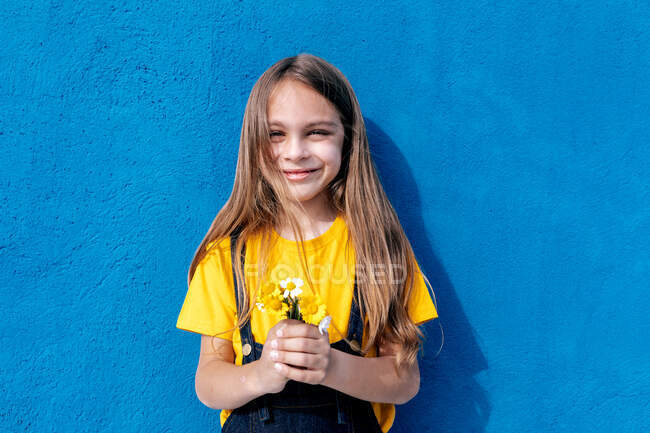 Adolescente segurando um monte de flores frescas enquanto estava no fundo da parede azul olhando para a câmera — Fotografia de Stock