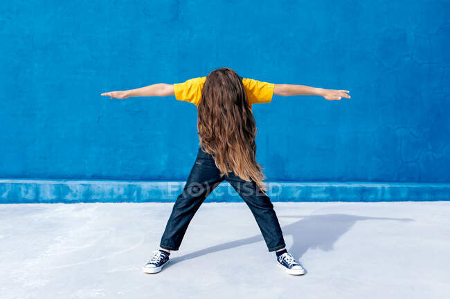 Adolescente enérgico anónimo en ropa fresca bailando con los brazos extendidos sobre fondo azul - foto de stock