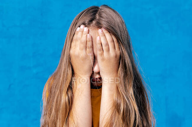 Невпізнавана сумна дитина прикриває обличчя, коли відчуває образи на синьому фоні в студії — стокове фото