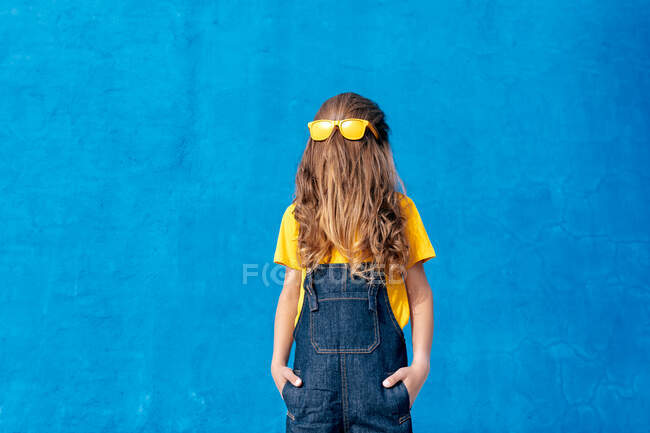 Anonymous cool teenager wearing yellow sunglasses on long hair covering face on blue background — Stock Photo
