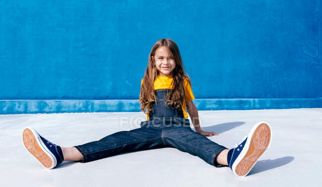 Позитивний підліток-хіпстер в прохолодному вбранні, сидячи на землі на тлі синьої стіни і дивлячись на камеру — стокове фото