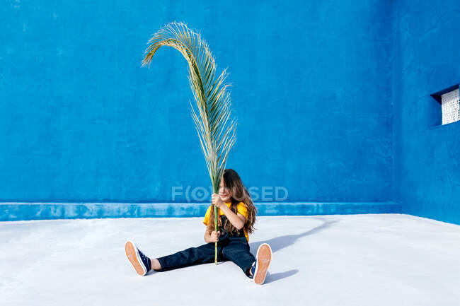 Adolescente sentado con enorme hoja de palmera en el fondo de la pared azul - foto de stock
