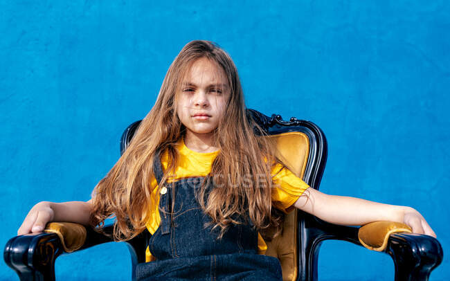 Nachdenklicher Teenager-Hipster sitzt auf Stuhl auf blauem Hintergrund und schaut in die Kamera — Stockfoto