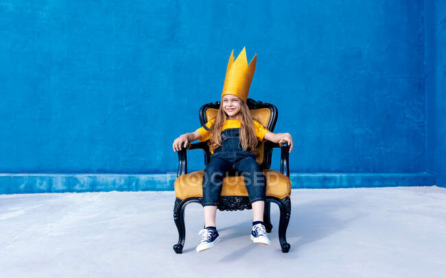 Contenu adolescent en couronne de papier doré assis sur le trône comme roi sur fond bleu regardant loin — Photo de stock