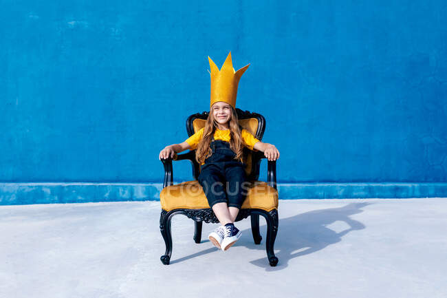 Contenu adolescent en couronne de papier doré assis sur le trône comme roi sur fond bleu regardant la caméra — Photo de stock