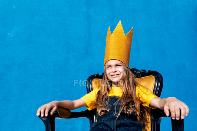 Зміст підлітка в золотій паперовій короні, що сидить на троні, як король на синьому фоні, дивлячись — стокове фото