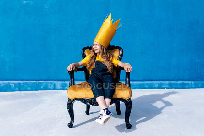 Adolescente contento con la corona de papel dorado sentado en el trono como rey sobre fondo azul mirando hacia otro lado - foto de stock